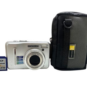 May include: A silver Samsung digital camera with a black carrying case. The camera has a zoom lens and a memory card slot. The memory card is a blue and yellow SD card labeled "Traveler 512MB".