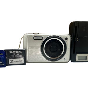 May include: A silver Samsung digital camera with a zoom lens, a black battery charger, a 2GB SD card, and a Samsung BP70A battery. The camera has a 5x zoom lens and the SD card is labeled "Traxdata Secure Digital".