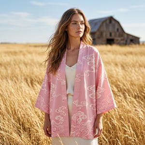 May include: A light pink open-front kimono jacket featuring a repeating white pattern of stylized clouds and waves. The garment has wide, three-quarter length sleeves and a straight, relaxed fit, worn over a simple, solid cream-colored camisole top.