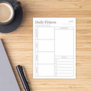 May include: A white Daily Fitness notepad with sections for breakfast, lunch, dinner, and snacks. It includes a workout section, a notes section, and a date line. A black pen and a cup of coffee are in the background.