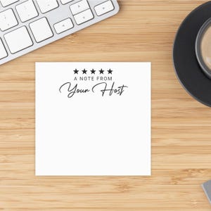 May include: A white square notepad with the text "A NOTE FROM Your Host" in black script. Above the text are five black stars. The notepad is on a wooden surface, with a keyboard and a coffee cup visible.
