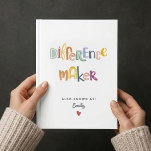 May include: A white hardcover book featuring the colorful text "Difference maker" in a playful font. Below this, the text reads "ALSO KNOWN AS: Emily" in a simple black font, followed by a small red heart icon centered at the bottom of the cover.
