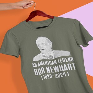 May include: Olive green t-shirt featuring a black and white portrait of Bob Newhart with the text "AN AMERICAN LEGEND BOB NEWHART (1929-2024)". The shirt is hanging on a wooden hanger.