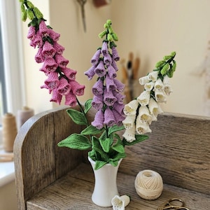 May include: A handcrafted display of crochet foxglove flowers in pink, purple, and white, arranged in a small white vase. The arrangement sits on a wooden shelf with a ball of yarn and scissors. The flowers have green leaves and stems.