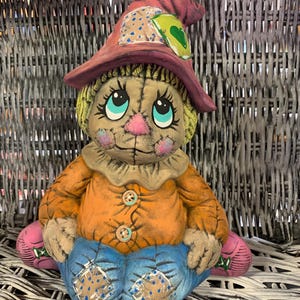 May include: A whimsical ceramic scarecrow figurine with a straw-like head and a wide-brimmed hat. The scarecrow wears an orange shirt with button details, blue denim pants, and pink foot coverings. The hat is adorned with patches.
