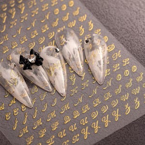 Puede incluir: Una lámina de pegatinas para uñas con el alfabeto en cursiva dorada. Cinco uñas acrílicas transparentes en forma de almendra están adornadas con letras doradas y un lazo negro con un brillante. El fondo es una superficie gris.