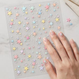Può includere: Un foglio trasparente di adesivi per unghie con una varietà di piccole stelle in tonalità pastello di rosa, giallo, blu e viola, intervallate da minuscoli puntini bianchi. Il testo PM YT4174 è stampato in piccole lettere nere sul foglio.