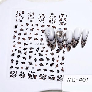 Puede incluir: Una hoja de pegatinas para uñas con un patrón de leopardo en marrón y negro y el texto MO-401. Junto a la hoja hay cuatro uñas postizas en forma de almendra con un degradado de blanco a marrón, puntas de leopardo y pequeños brillantes.
