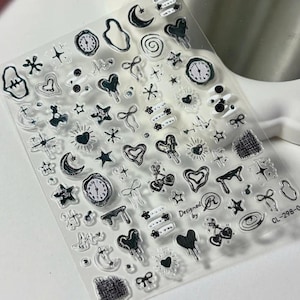 Può includere: Un foglio trasparente di adesivi per unghie con illustrazioni a inchiostro nero di cuori, orologi, stelle, fiocchi, lune, ciliegie e spirali. Il foglio include il testo Designed by R e il codice prodotto CL-298-0 stampato in nero.