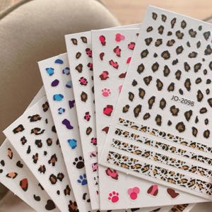 Puede incluir: Una colección de hojas de pegatinas blancas para uñas con varios patrones de leopardo en negro, marrón, azul, rosa y rojo. La hoja superior está etiquetada como JO-2098 e incluye varias tiras horizontales con diseños de estampado de leopardo.