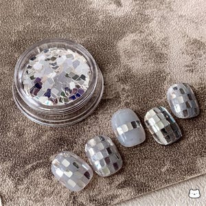 May include: A clear plastic jar filled with small, square, reflective silver nail art sequins, accompanied by four press-on nail tips decorated with a grid of these same metallic silver squares arranged in a mosaic pattern on a light gray base.