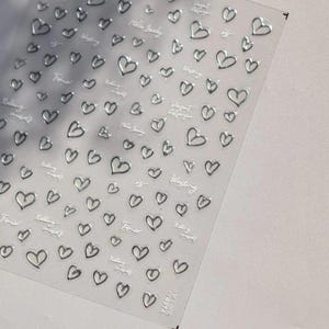 May include: A clear sheet of adhesive nail stickers featuring numerous small silver heart outlines and cursive text phrases including Hello Lovely, Rolling myself, and Time is wasting. The stickers are arranged in a grid pattern on a transparent backing.
