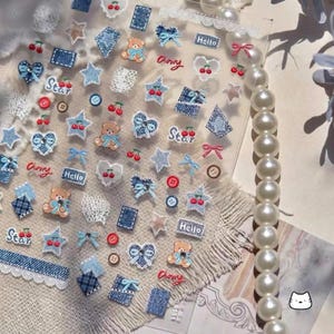 May include: A sheet of adhesive stickers featuring various designs including teddy bears with blue bows, denim-textured hearts, stars, cherries, red and brown buttons, pink bows, white lace patterns, and rectangular labels with the text Hello and Star.