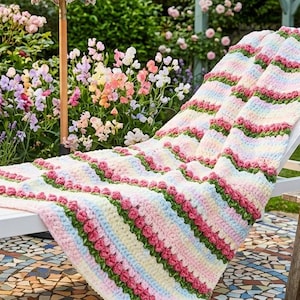 Op de afbeelding: Een gestreepte, gehaakte deken in roze, wit en groen ligt over een witte ligstoel. De scène speelt zich af in een tuin met een bloemenparasol, bloemen en een kleine tafel met een kan fruit.