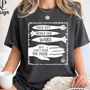 May include: Dark grey t-shirt with a white graphic that reads "THEY GOT MONEY FOR WARS BUT CAN'T FEED THE POOR." The shirt is a Comfort Colors brand in the color "Pepper."