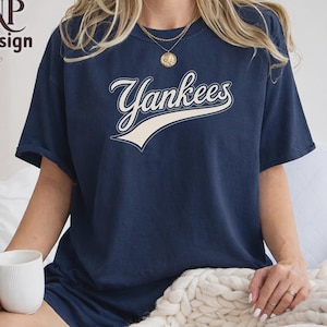 May include: A navy blue short-sleeve t-shirt featuring the word Yankees in a white script font with a swooping underline. The shirt has a crew neckline and is made of a solid-colored fabric. The design is centered on the chest area of the garment.