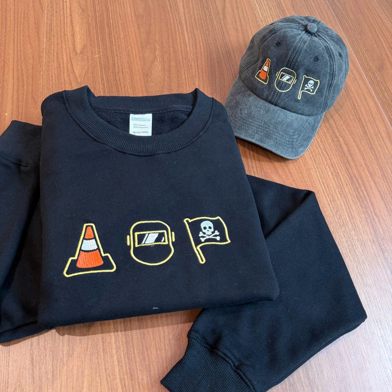 Peut inclure: Un sweat-shirt noir et une casquette de baseball grise. Le sweat-shirt pr&eacute;sente des images brod&eacute;es d'un c&ocirc;ne de signalisation, d'un masque de soudure et d'un drapeau avec une t&ecirc;te de mort. La casquette a les m&ecirc;mes images brod&eacute;es.