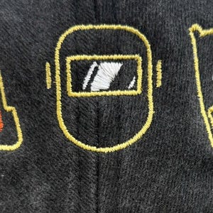 Peut inclure: Tissu gris fonc&eacute; avec des contours brod&eacute;s en jaune. Le motif comprend un c&ocirc;ne de signalisation, un masque de soudeur et un drapeau avec une t&ecirc;te de mort et des os crois&eacute;s. La broderie est orange, blanche et jaune.