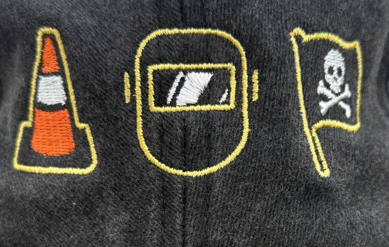 Peut inclure: Tissu gris fonc&eacute; avec des contours brod&eacute;s en jaune. Le motif comprend un c&ocirc;ne de signalisation, un masque de soudeur et un drapeau avec une t&ecirc;te de mort et des os crois&eacute;s. La broderie est orange, blanche et jaune.