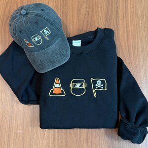Peut inclure: Un sweat-shirt noir et une casquette de baseball en jean gris. Le sweat-shirt pr&eacute;sente des images brod&eacute;es d'un c&ocirc;ne de signalisation, d'un masque de soudure et d'un drapeau avec une t&ecirc;te de mort. La casquette a les m&ecirc;mes motifs brod&eacute;s.
