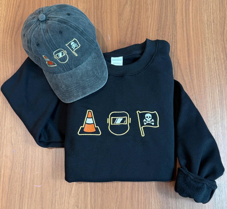 Peut inclure: Un sweat-shirt noir et une casquette de baseball en jean gris. Le sweat-shirt pr&eacute;sente des images brod&eacute;es d'un c&ocirc;ne de signalisation, d'un masque de soudure et d'un drapeau avec une t&ecirc;te de mort. La casquette a les m&ecirc;mes motifs brod&eacute;s.