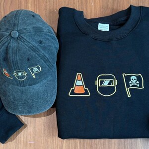 Peut inclure: Une casquette de baseball gris fonc&eacute; et un sweat-shirt noir. La casquette pr&eacute;sente des graphiques brod&eacute;s orange et blancs. Le sweat-shirt pr&eacute;sente des images brod&eacute;es d'un c&ocirc;ne de signalisation, d'un masque de soudure et d'un drapeau avec une t&ecirc;te de mort.