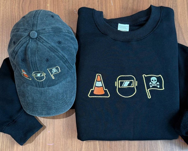 Peut inclure: Une casquette de baseball gris fonc&eacute; et un sweat-shirt noir. La casquette pr&eacute;sente des graphiques brod&eacute;s orange et blancs. Le sweat-shirt pr&eacute;sente des images brod&eacute;es d'un c&ocirc;ne de signalisation, d'un masque de soudure et d'un drapeau avec une t&ecirc;te de mort.