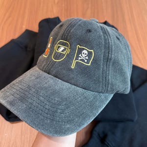 Peut inclure: Casquette de baseball grise avec des motifs brod&eacute;s jaunes et oranges. Les motifs comprennent un c&ocirc;ne de signalisation, un masque de soudeur et un drapeau t&ecirc;te de mort. La casquette est faite d'une mati&egrave;re douce et us&eacute;e.