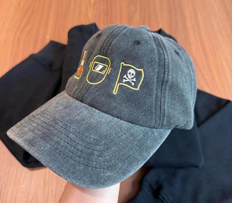Peut inclure: Casquette de baseball grise avec des motifs brod&eacute;s jaunes et oranges. Les motifs comprennent un c&ocirc;ne de signalisation, un masque de soudeur et un drapeau t&ecirc;te de mort. La casquette est faite d'une mati&egrave;re douce et us&eacute;e.