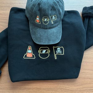 Peut inclure: Un sweat-shirt noir et une casquette de baseball avec les lettres "AOP" et des graphiques. Les motifs comprennent un c&ocirc;ne de signalisation, un masque de soudure et un drapeau de pirate. Les deux articles sont gris fonc&eacute;.