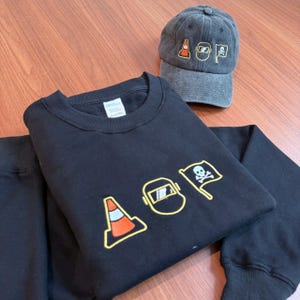 Peut inclure: Sweat-shirt noir et casquette grise avec des motifs brod&eacute;s. Le sweat-shirt pr&eacute;sente un c&ocirc;ne de signalisation, un masque de soudure et un drapeau t&ecirc;te de mort avec les lettres "AOP". La casquette a un design similaire.