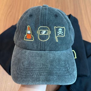 Peut inclure: Casquette de baseball grise avec des graphiques brod&eacute;s jaunes et oranges. Les graphiques comprennent un c&ocirc;ne de signalisation, un masque de soudure et un drapeau avec une t&ecirc;te de mort. La casquette est en tissu doux et us&eacute;.
