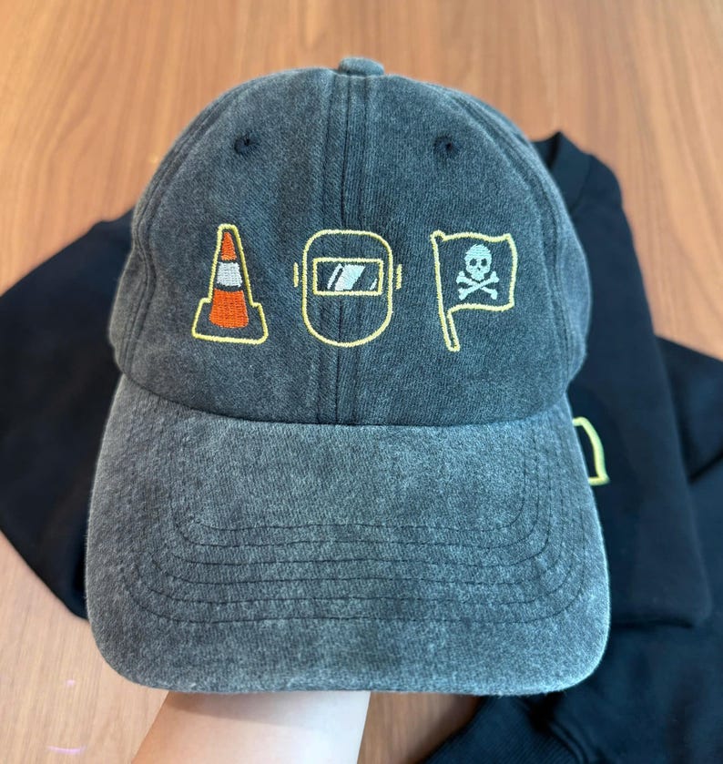 Peut inclure: Casquette de baseball grise avec des graphiques brod&eacute;s jaunes et oranges. Les graphiques comprennent un c&ocirc;ne de signalisation, un masque de soudure et un drapeau avec une t&ecirc;te de mort. La casquette est en tissu doux et us&eacute;.