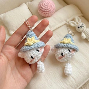 May include: Crocheted cloud-shaped keychains with blue hats and yellow stars. The white clouds have rosy cheeks and small black eyes. Each keychain has a silver chain and a small white dangling piece. A pink crocheted ball and a white crocheted bunny are in the background.