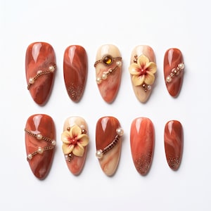 May include: A set of ten almond-shaped artificial nails in shades of brown, beige, and orange. Some nails feature decorative chains, pearls, and 3D flower accents. A few nails have glitter at the tips.