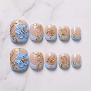 May include: A set of ten press-on nails featuring a light blue and nude gradient base. The design includes raised 3D light blue flowers with crystal centers, gold filigree swirls, gold letter AB and D accents, and small gold leaf patterns.