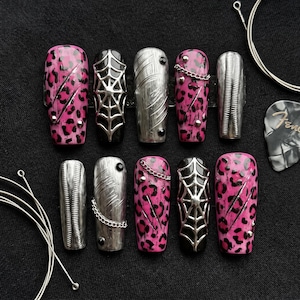May include: A set of ten press-on nails featuring a mix of designs including hot pink leopard print, black spider webs, metallic silver textures, and small silver chains. The nails are arranged in two rows alongside guitar strings and a guitar pick.
