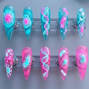 May include: Ten stiletto-shaped press-on nails featuring scientific motifs. The top row has translucent teal bases with pink cell and DNA designs. The bottom row has translucent pink bases with teal cell and DNA designs, all showcasing raised 3D art.