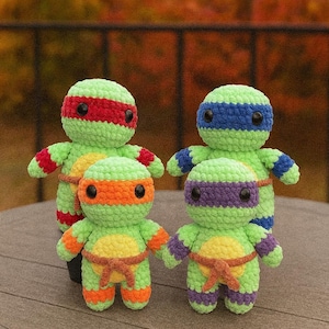 May include: Four handmade crochet figures of the Teenage Mutant Ninja Turtles. Each figure is green with a different coloured mask: red, blue, orange, and purple. They have brown belts and black eyes. The figures are standing on a brown surface.