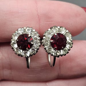 Może przedstawiać: Para srebrnych kolczyków na klipsie z czerwonym kryształem otoczonym mniejszymi, przezroczystymi kryształami.