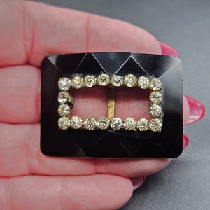 May include: A black plastic buckle with a rectangular frame of clear rhinestones. The buckle is designed to be used for a belt or strap.