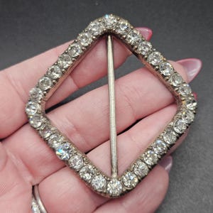 May include: A vintage, diamond-shaped buckle with a silver finish. The buckle is encrusted with numerous clear, sparkling rhinestones. A silver bar runs through the center of the buckle. The buckle is held in a hand.