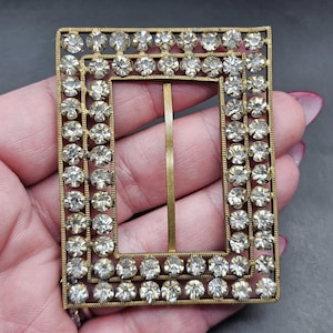 May include: A gold-toned metal buckle with a rectangular frame and a center bar. The frame is decorated with a border of clear rhinestones.