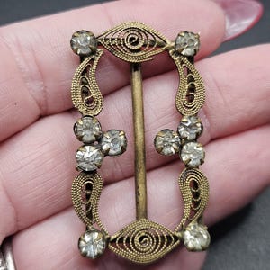 May include: Vintage brass buckle with clear rhinestone detailing. The buckle has an open filigree design with decorative swirls and a central bar. The rhinestones are round. A classic accessory.