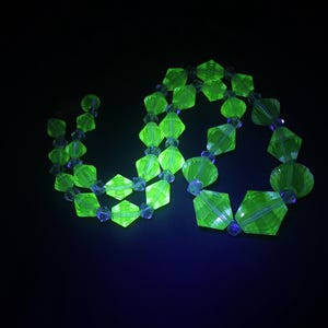 Pode incluir: Um colar verde néon com contas geométricas facetadas e contas azuis redondas menores. O colar é exibido em um fundo preto, destacando sua aparência brilhante e brilhante. As contas têm uma qualidade transparente.