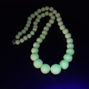 Puede incluir: Un collar compuesto por cuentas lisas, redondas y de color verde lima pálido que aumentan de tamaño hacia el centro. Las cuentas exhiben una cualidad luminosa y brillante sobre el fondo oscuro, y el collar se asegura con un pequeño broche metálico.