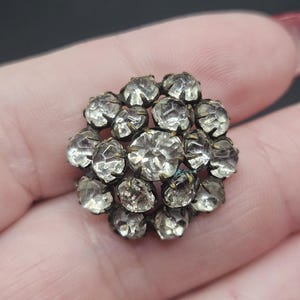 Pode incluir: Um broche circular com um grupo de pedras transparentes facetadas engastadas numa armação de metal escuro e envelhecido. As pedras estão dispostas num padrão denso e multicamadas com uma pedra central maior rodeada por pedras mais pequenas.