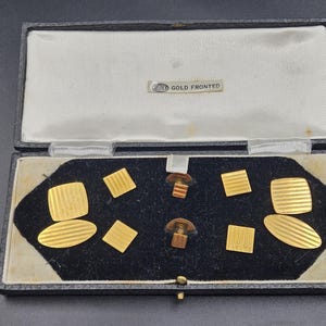 May include: A black box with a white interior and a black velvet base, holding gold-coloured cufflinks and shirt studs. The cufflinks and studs are square and oval, with striped and solid patterns. The box has a label reading "GOLD FRONTED".