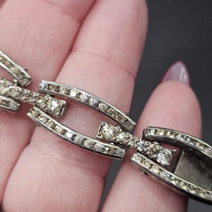 Puede incluir: Primer plano de una pulsera plateada con un diseño de eslabones. Cada eslabón está adornado con pequeños y brillantes pedrería. La pulsera se sostiene sobre un fondo neutro, resaltando sus intrincados detalles y su estilo vintage.