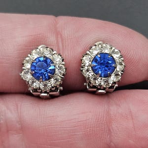 Könnte beinhalten: Ein Paar Vintage-Clip-Ohrringe. Jeder Ohrring hat einen großen, runden, tiefblauen Edelstein, der von einem Kranz aus kleineren, klaren Kristallen umgeben ist. Die Ohrringe haben eine silberfarbene Metalleinfassung und einen Clipverschluss.